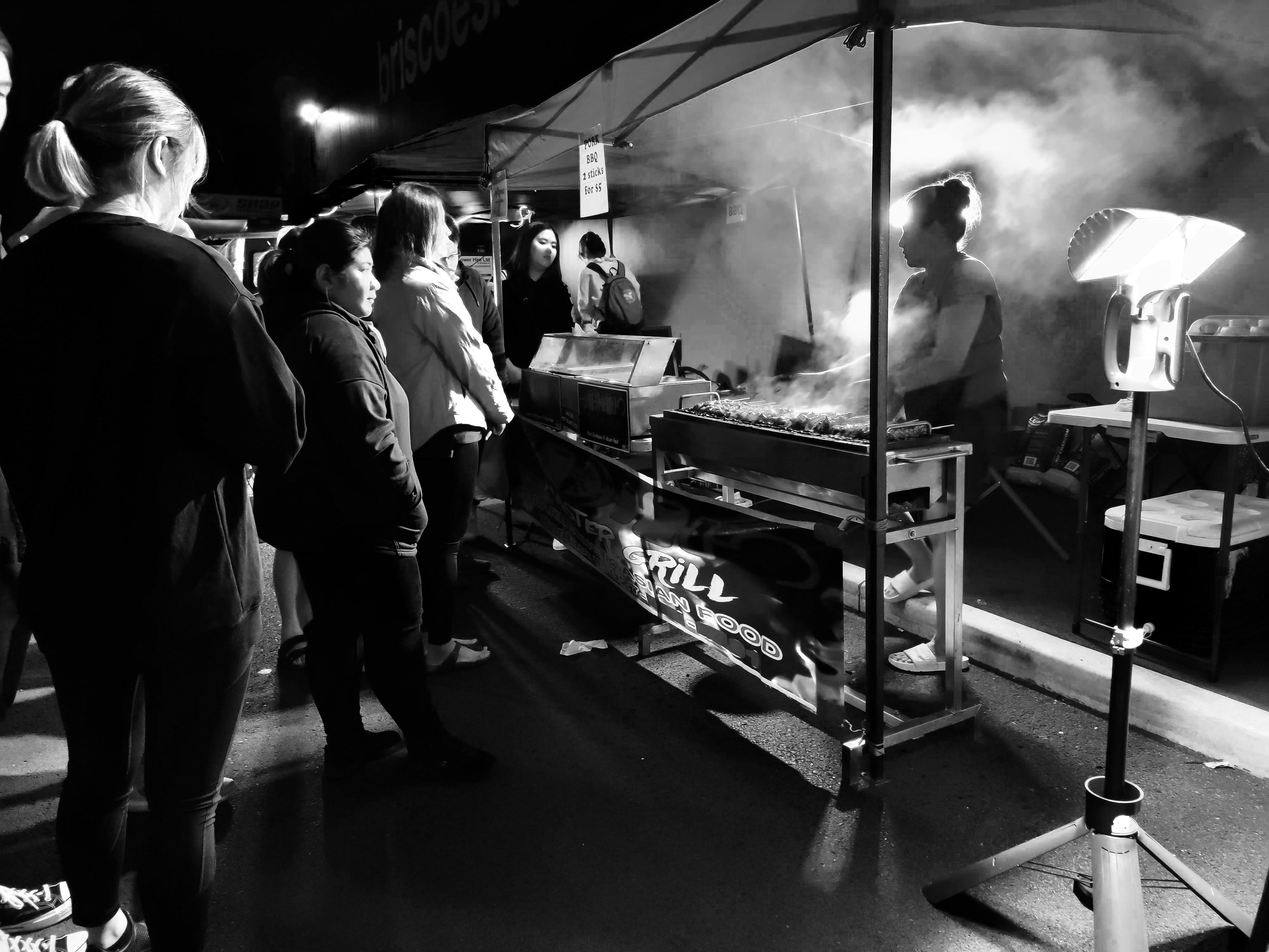 People queuing to buy meat skewers from a woman cooking them under a gazebo. There is a strong electric light creating high contrast, Christchurch.