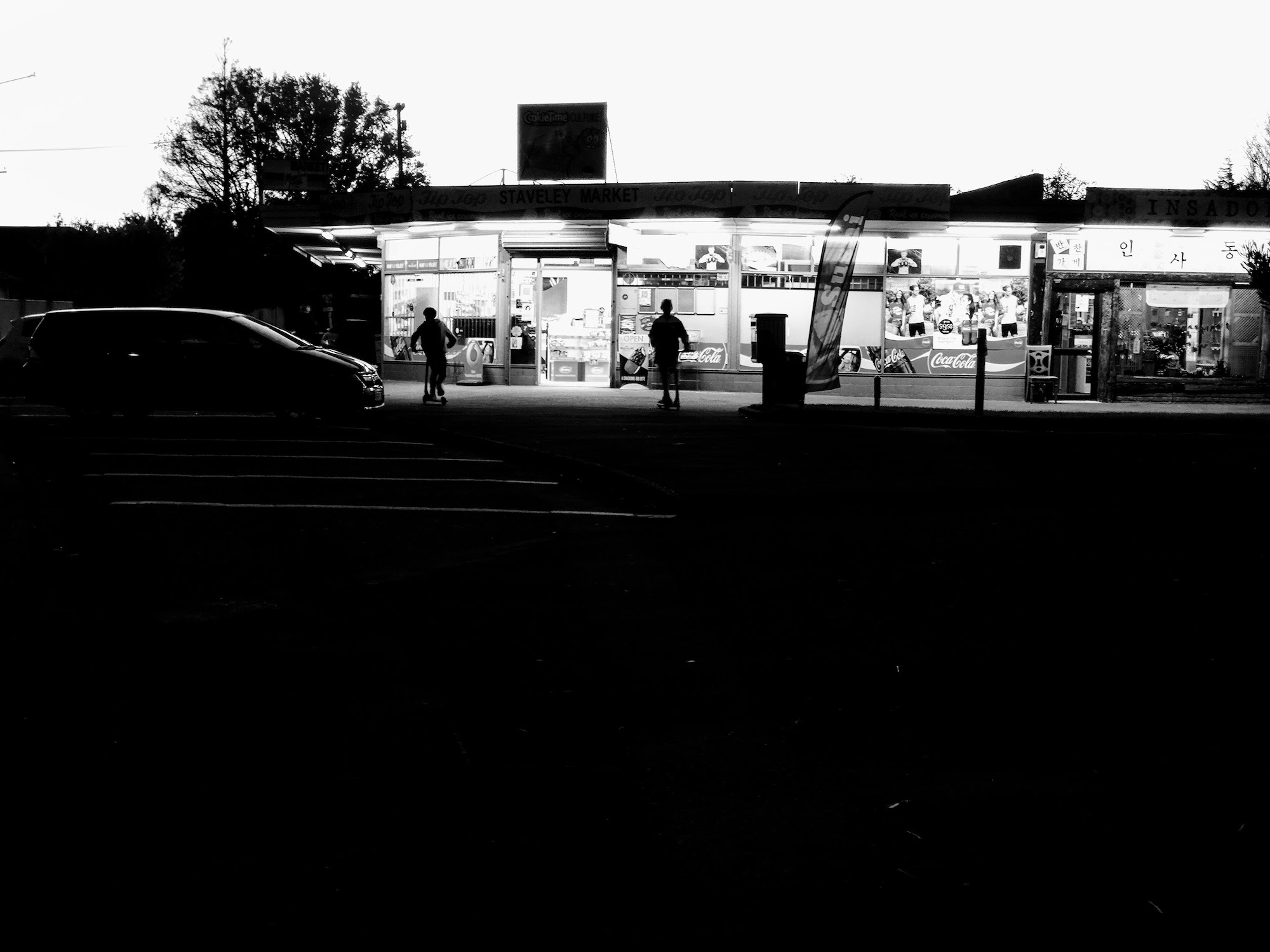 Dark foreground with light sky, dairy lights on and a couple of kids playing with their scooters.