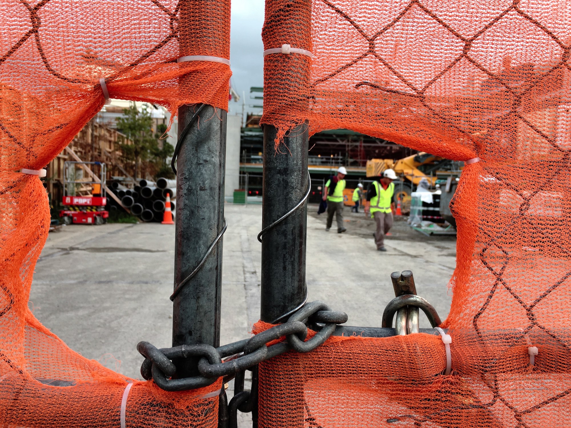 Workers wearing high visibility vests behind a fence covered with orange netting + Velvia 50 film simulation, Christchurch.