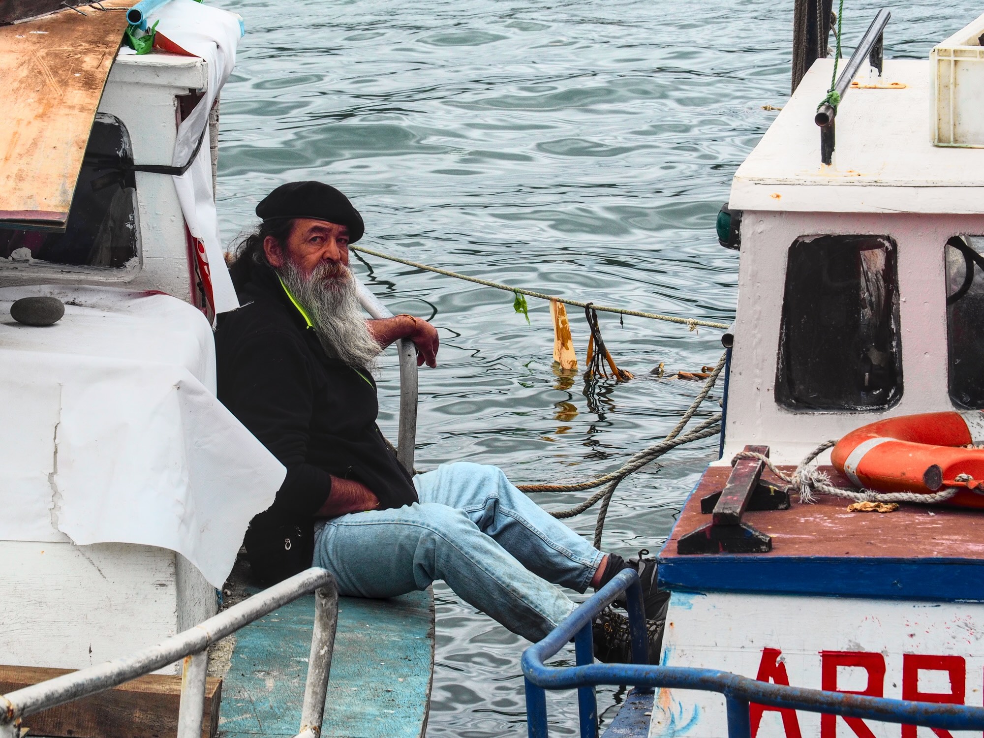 Long bearded fisherman sitting on a boat with his feet touching another boat, Dalcahue.