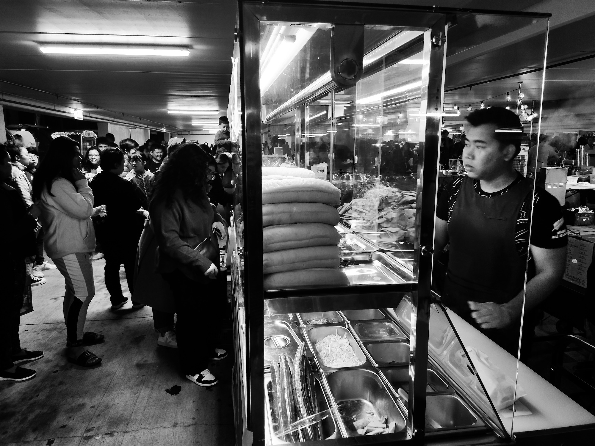 Background: people queuing to but food. Foreground: man behind food display, Christchurch.