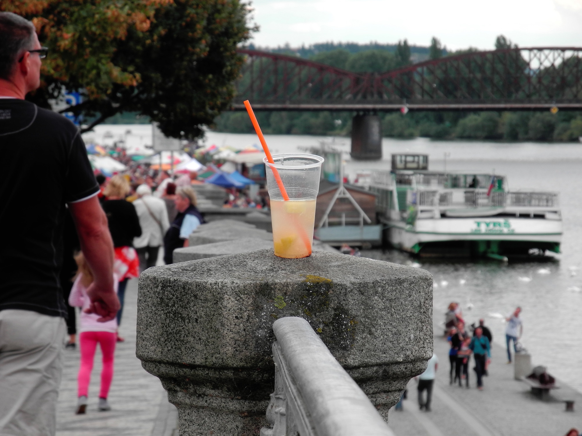 Half-drunk juice glass with an orange straw, resting on a stone edge next to a river market + Fuji Velvia 50 simulation, Prague.