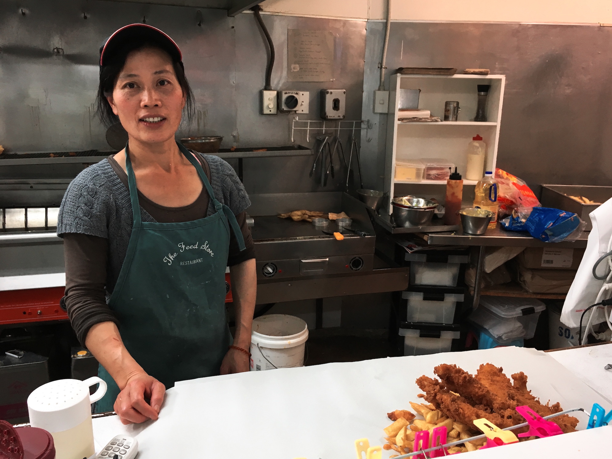 Woman looking directly to the camera before wrapping 3 crumbed fish and 2 scoops of chips. Fryer and grill on the back + Fuji Provia 100F simulation, Christchurch.