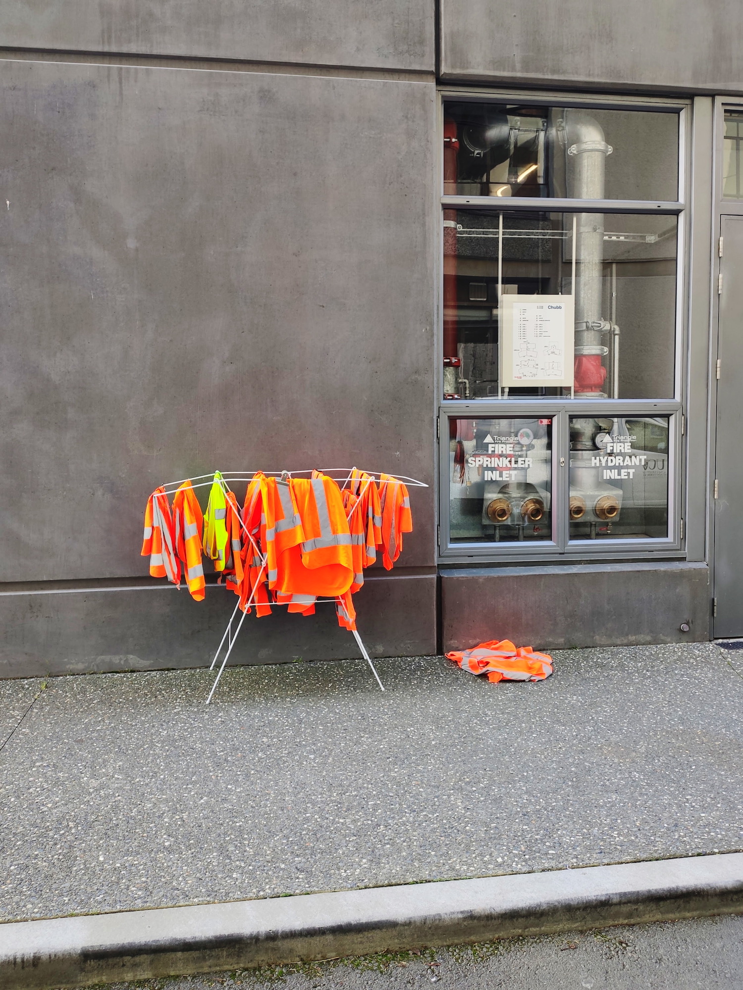 Over ten high visibility vests, all orange but a yellow one, hanging on a clothes drier. A window next to them shows a number of big pipes + Velvia 100 simulation, Christchurch.